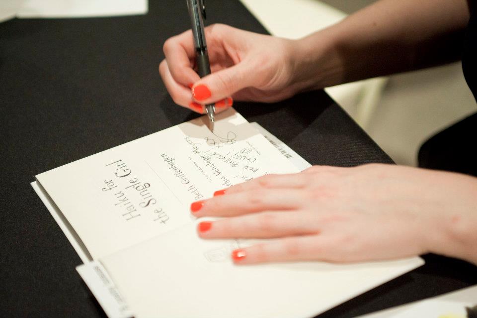 Photograph of the book "Haiku for the Single Girl" on a desk, opened to the title page, and with a pair of hands (the author Beth Griffenhagen) on the book, one hand holding the book open, the other autographing it